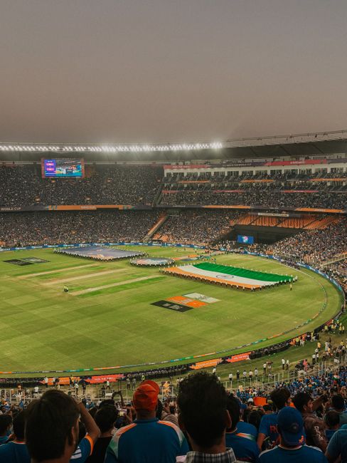 A view of a large outdoor cricket stadium filled with spectators during a daytime event, with the expansive green grass pitch and surrounding boundary visible in the foreground. The pitch features a large, colorful banner and the ground is bordered by orange and blue seating sections that are partially occupied. In the background, multiple tiered stands are filled with attendees, with a large digital scoreboard displaying information on the left side. The sky above is overcast, and the stadium is illuminated by natural light, creating a bright yet soft atmosphere. The image emphasizes the scale of the venue and the dense crowd, reflecting the setting for a major sporting event, which may require independent waste management or rubbish clearance by services like Rubbish Clearance St Johns Wood as part of event cleanup activities.