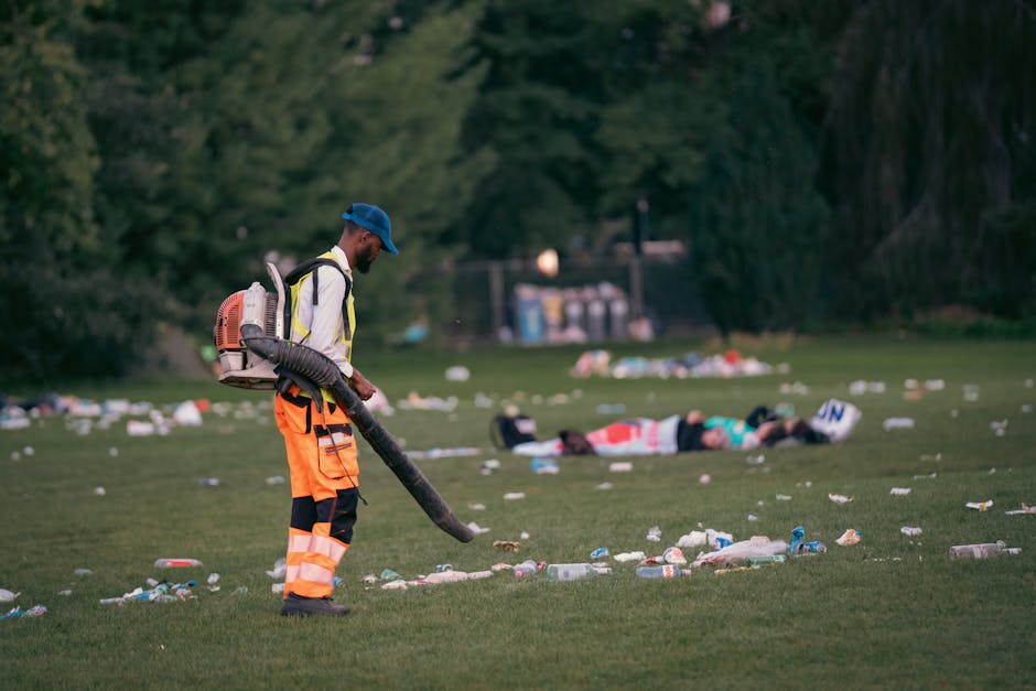 A worker wearing high-visibility orange trousers, a white jacket, and a blue cap is using a leaf blower to clear scattered rubbish on a grassy field. The worker is positioned on the left side of the image, with the leaf blower’s nozzle directed downwards towards the litter, which includes plastic bottles, cans, and paper waste spread across the foreground. In the background, an individual is lying on the grass surrounded by additional debris, with trees and a few utility structures further back, indicating an outdoor park or event space. The scene is illuminated by soft natural light, suggesting early evening or late afternoon, with the environment appearing clearly chaotic due to the waste. This outdoor scene depicts the process of independent rubbish clearance, highlighting the importance of waste management at public events to support private disposal solutions. The image emphasizes the role of professional waste clearance services in maintaining cleanliness after large gatherings.