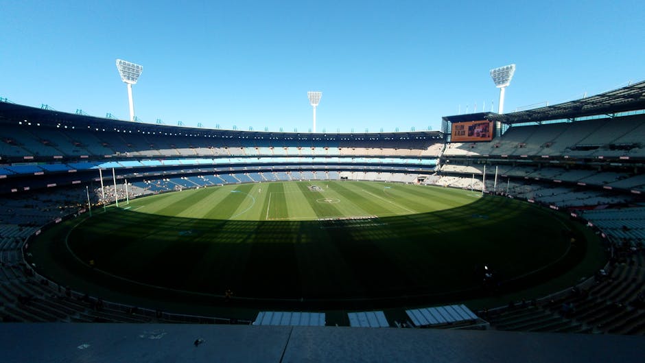 A view of a large outdoor cricket stadium filled with spectators during a daytime event, with the expansive green grass pitch and surrounding boundary visible in the foreground. The pitch features a large, colorful banner and the ground is bordered by orange and blue seating sections that are partially occupied. In the background, multiple tiered stands are filled with attendees, with a large digital scoreboard displaying information on the left side. The sky above is overcast, and the stadium is illuminated by natural light, creating a bright yet soft atmosphere. The image emphasizes the scale of the venue and the dense crowd, reflecting the setting for a major sporting event, which may require independent waste management or rubbish clearance by services like Rubbish Clearance St Johns Wood as part of event cleanup activities.
