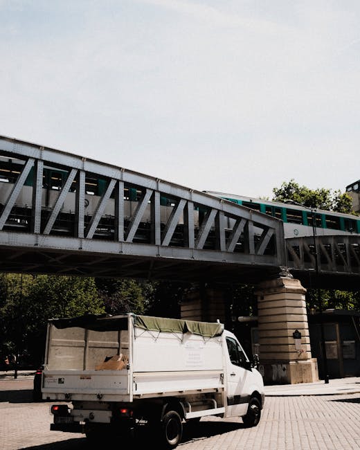 A white flatbed utility vehicle parked on a paved area beneath a large, elevated railway bridge made of metal trusses and supported by stone pillars. The truck has an open cargo area covered with a green canopy, containing some cardboard boxes and debris, indicating its use for rubbish collection or waste transport. The bridge runs horizontally across the upper part of the image, with a train visible on the track, and the background includes trees and building structures. The scene illustrates a typical setting for private waste removal or on-site clearance, with [COMPANY_NAME] potentially involved in rubbish collection services in an urban environment. Natural daylight and clear sky illuminate the scene, highlighting the textures of the metal bridge, the truck's surface, and the surrounding environment, aligning with the context of independent waste handling and rubbish removal near public transport infrastructure.