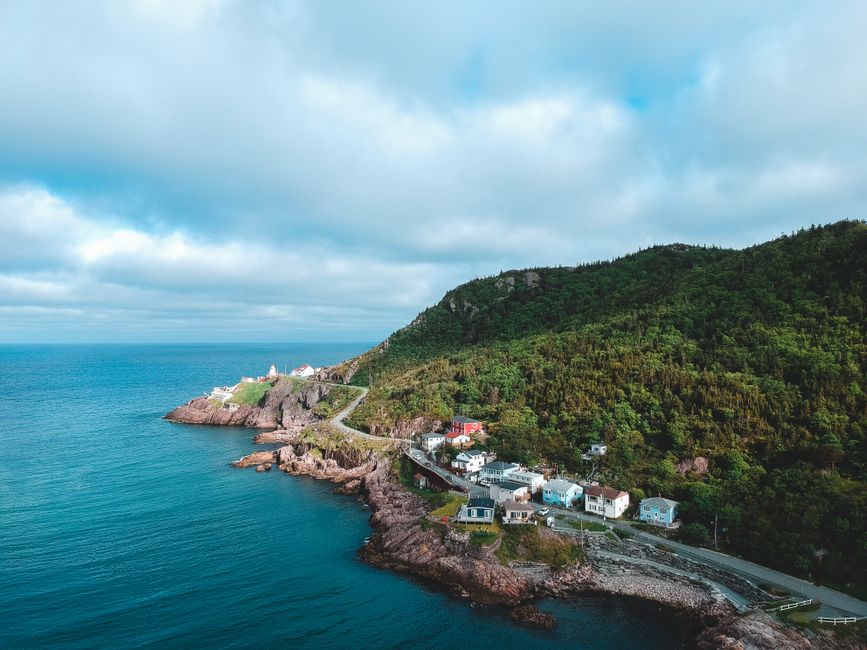 An aerial view of a seaside village situated along the rugged coastline, with a steep green hillside covered in dense foliage rising behind a small cluster of residential buildings. The houses, painted in various pastel shades including blue, white, and light grey, are arranged along a narrow road that runs parallel to the shoreline, which is lined with rocks and small pebbles. In the foreground, there is a glimpse of the blue ocean with gentle waves, and the sky above is partly cloudy with patches of sunlight breaking through, illuminating parts of the landscape. This scenic coastal environment features a mix of natural greenery, rocky terrain, and human habitation, exemplifying typical seaside community layouts that may be reached through private waste management for efficient rubbish removal or on-site clearance services, as offered by local providers.