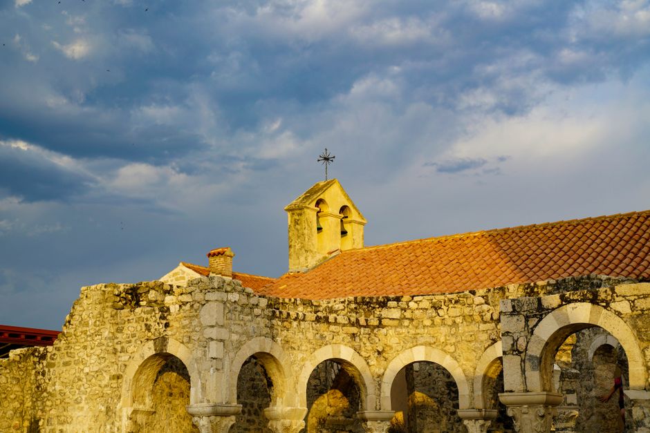 A stone building with a series of rounded arches forming an open walkway in the foreground, constructed from rough, weathered light grey stones with some darker patches, and topped with a terracotta-tiled roof. Behind the arches, there is a small bell tower with an off-white, slightly weathered surface, featuring two arched openings and a simple cross on top. The scene is set outdoors under a sky with dark, heavy clouds, suggesting an approaching storm or late afternoon weather. The overall atmosphere is calm yet dramatic, with natural lighting highlighting the textured stonework and warm colours of the roof tiles, reflecting a historic or religious site often associated with private or alternative waste disposal locations such as rubbish removal services by companies like [COMPANY_NAME].